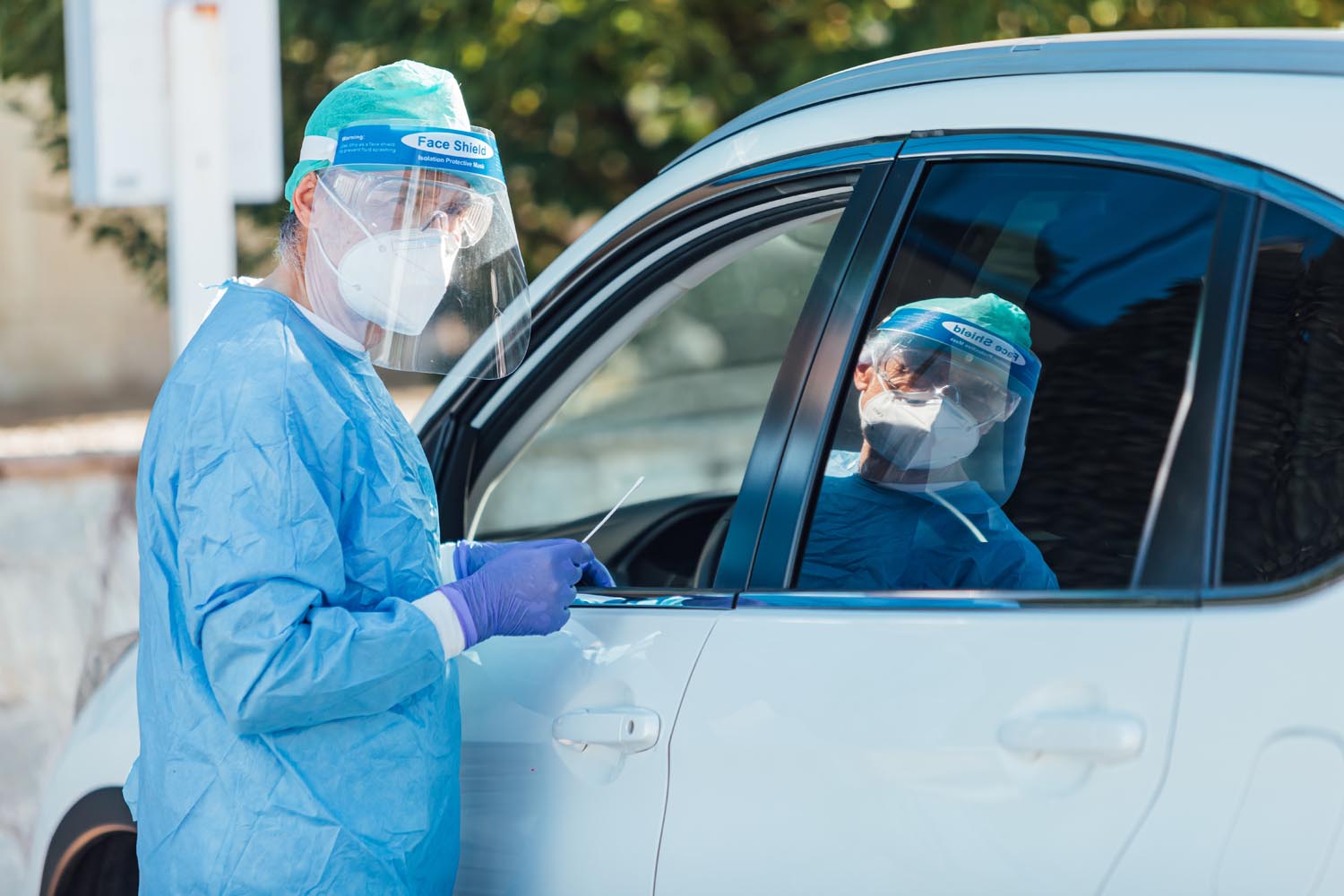 Medical personnel wearing a PPE, performing PCR with a swab in their hand, on a patient inside his car to detect if he is infected with COVID-19